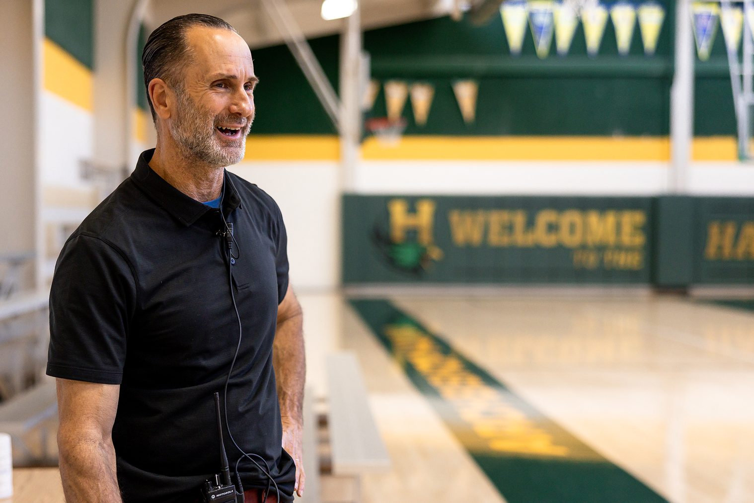 Coach or staff member standing in gymnasium with ‘Welcome Hawks’ signage.